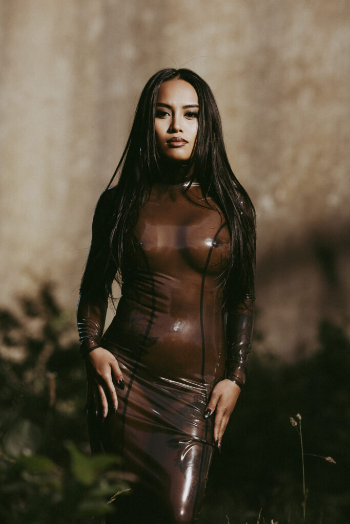 MissThaiTiny wearing a translucent brown latex dress by Simon O., photographed by Lasse Lagoni glossy, confident, and elegant fashion portrait captured outdoors in warm light.