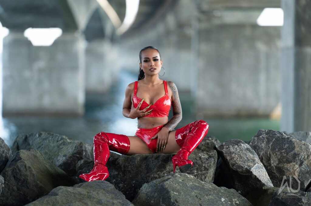 Miss ThaiTiny posing under a bridge in a glossy red latex outfit and thigh-high boots, photographed by Arsumbrarum.
