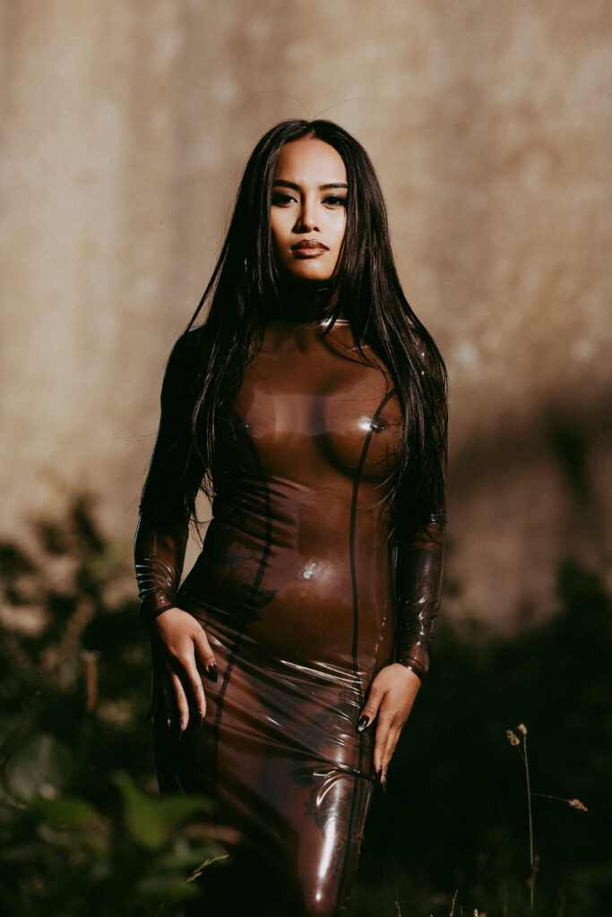 Miss ThaiTiny posing outdoors in a sheer brown latex dress under golden sunlight, photographed by Lasse Lagoni.