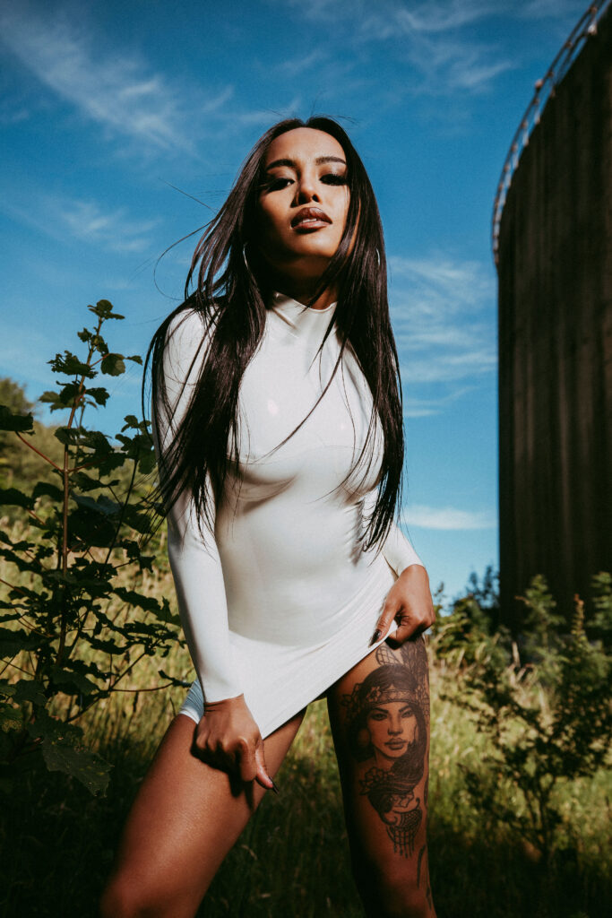 Miss ThaiTiny wearing a white latex dress outdoors under a bright blue sky, photographed by Lasse Lagoni.
